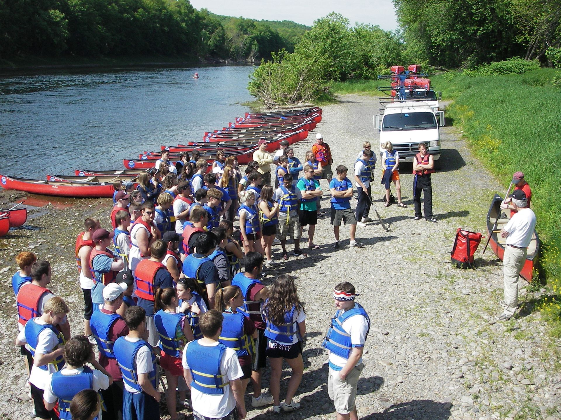 A group of people wearing life jackets are standing in front of a river with adventure sports canoes