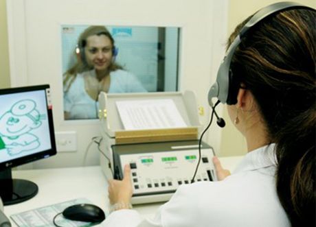 Una mujer con auriculares se somete a una prueba de audición en una cabina insonorizada. Un audiólogo opera el equipo de prueba, mirando un monitor de computadora.