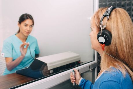 Mujer con auriculares sometiéndose a una prueba de audición en una cabina insonorizada, mientras un trabajador sanitario ajusta el equipo.