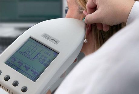 Un profesional médico realiza un examen de oído a un paciente con un dispositivo portátil. El dispositivo muestra una gráfica.