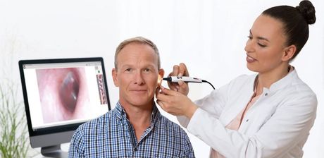 Un médico examina la oreja de un hombre con un otoscopio, una imagen digital del oído se muestra en una pantalla de computadora.