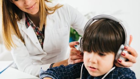 Una mujer con bata blanca ayuda a un niño a ponerse los auriculares para una prueba de audición. El lugar parece ser un consultorio médico.
