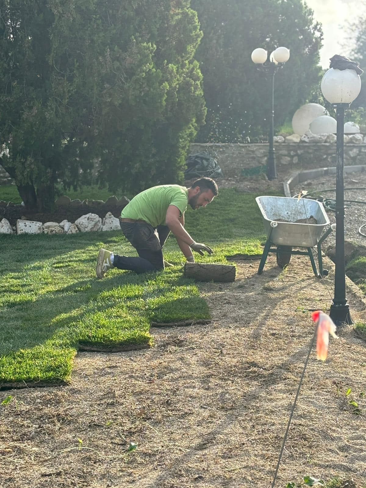 Uomo che stende il prato con una carriola nelle vicinanze. Camicia verde e jeans. All'aperto, giornata di sole.
