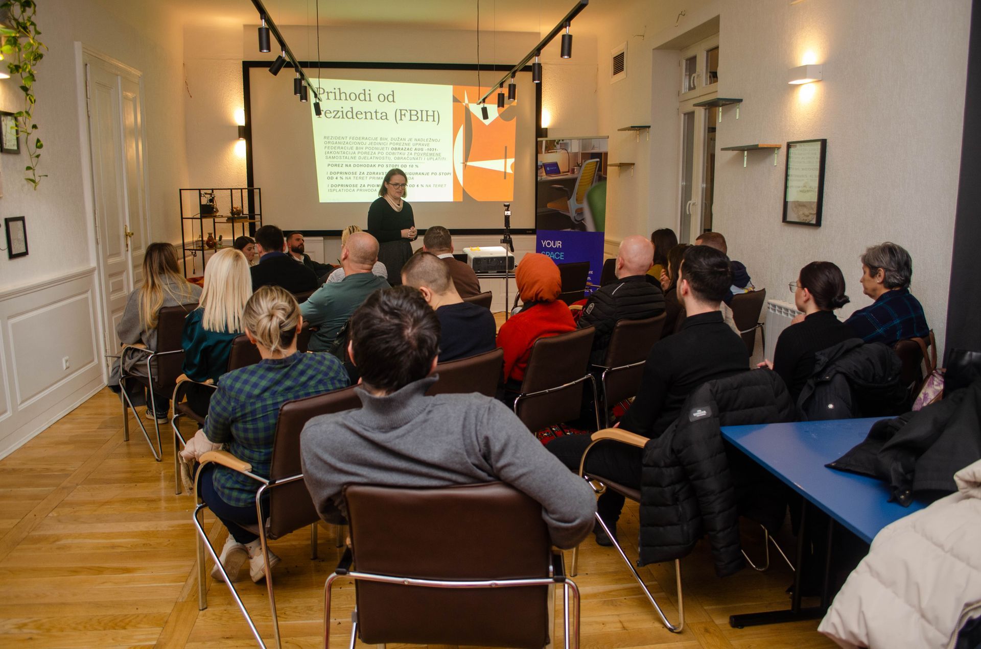 A woman giving a presentation to a seated audience in a meeting room.