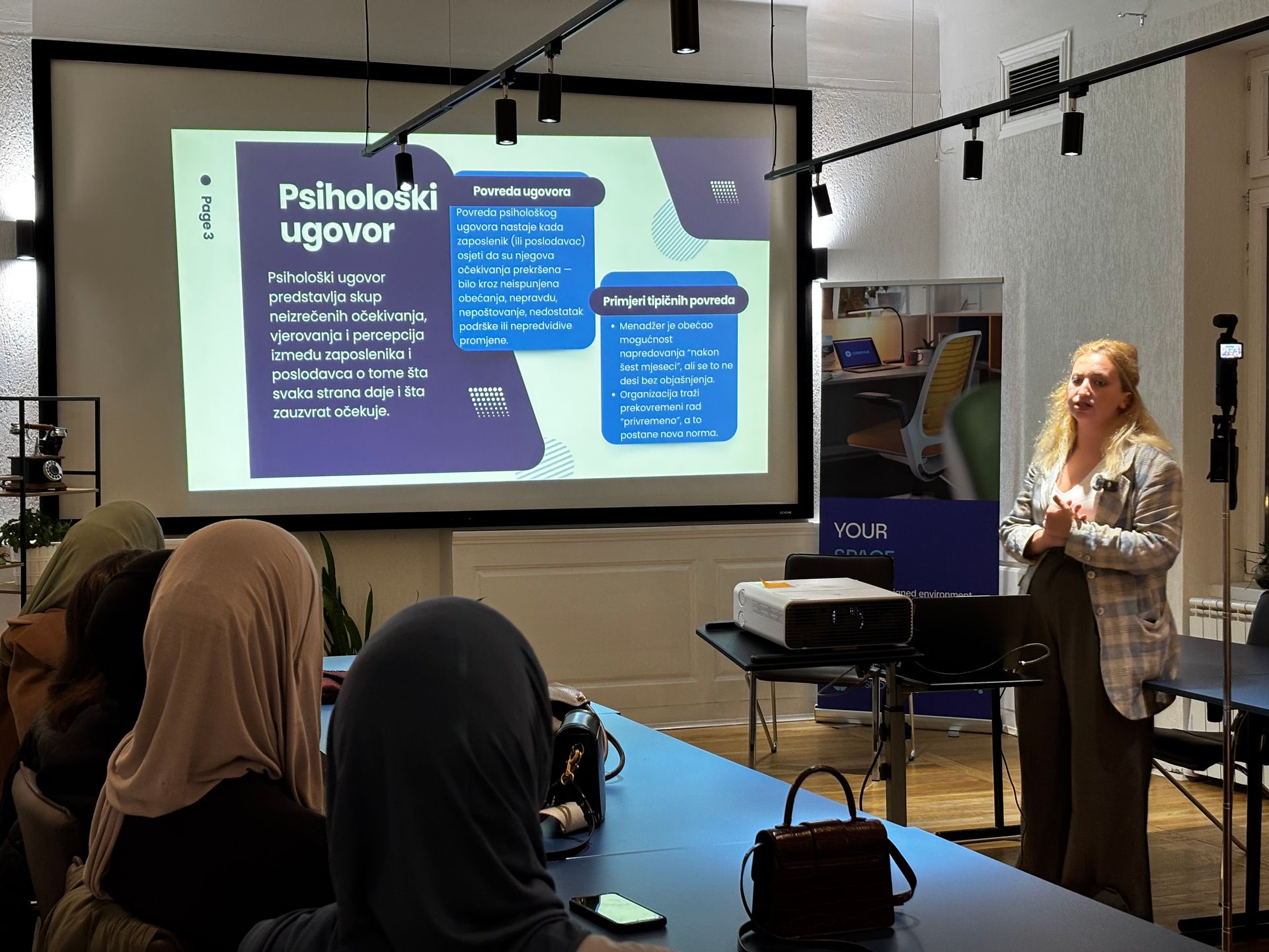 A woman presents to a seated audience with a projector screen showing text.
