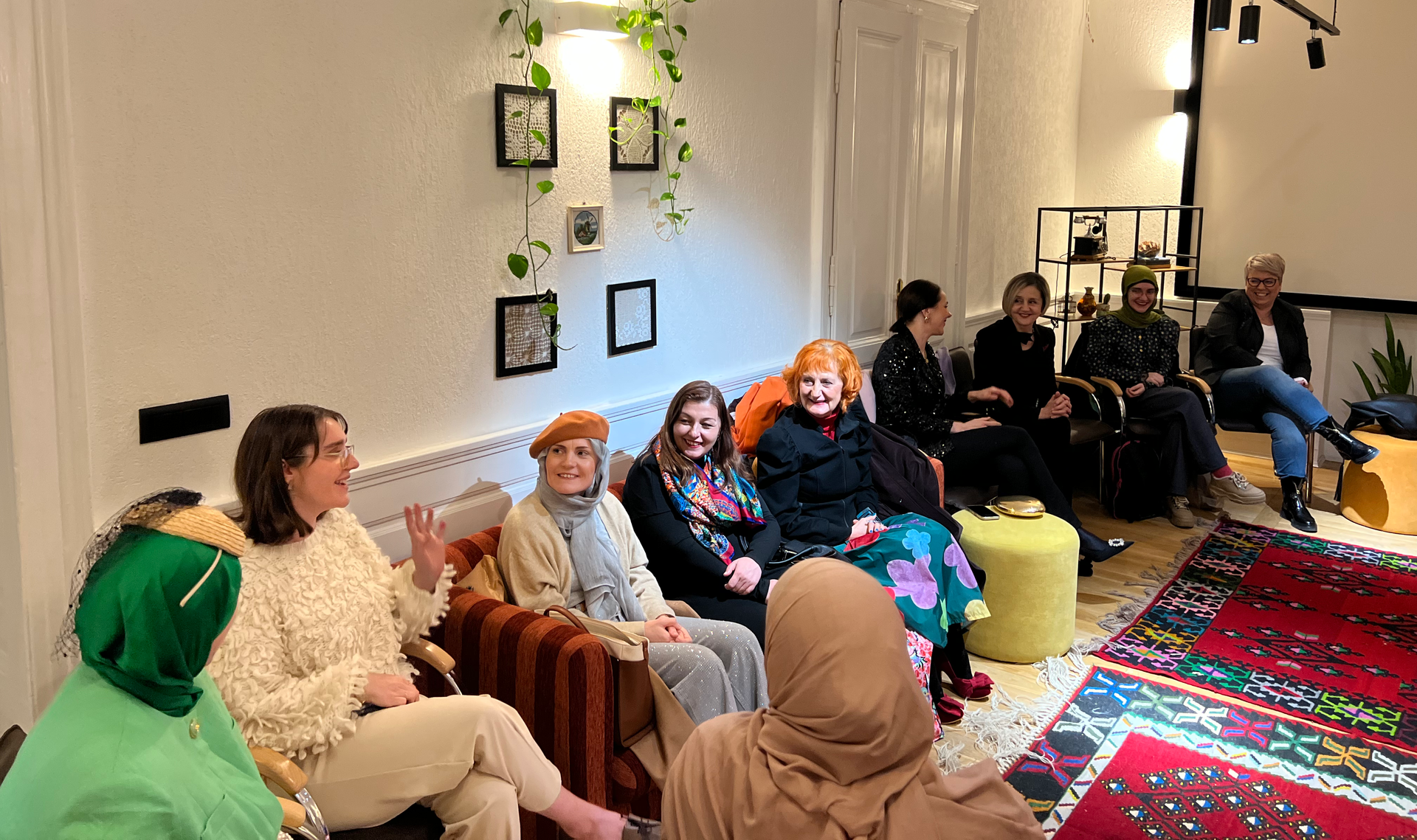 A group of women sit and converse in a bright room. Some are on a couch, others in chairs. There's a decorative rug and plants.