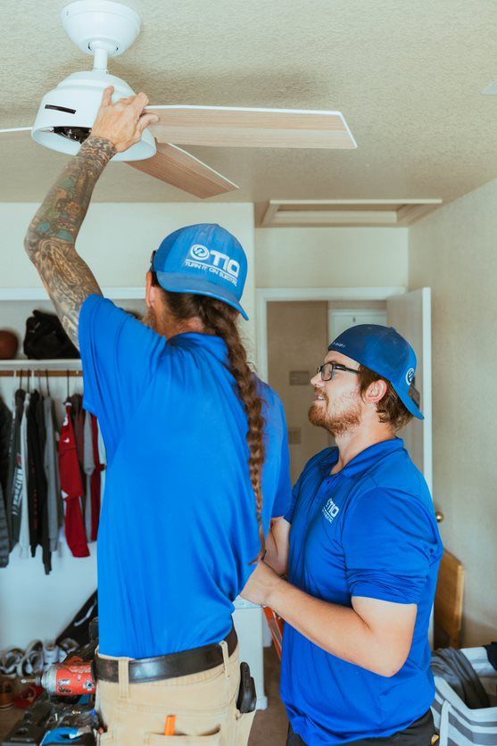 Two men are working on a ceiling fan in a bedroom.