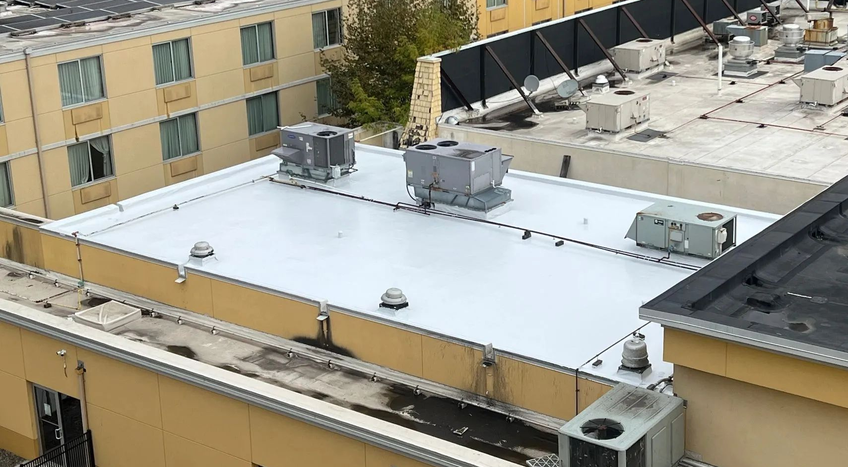 An aerial view of a flat, white-coated commercial roof with HVAC units and skylights, surrounded by tan building walls.