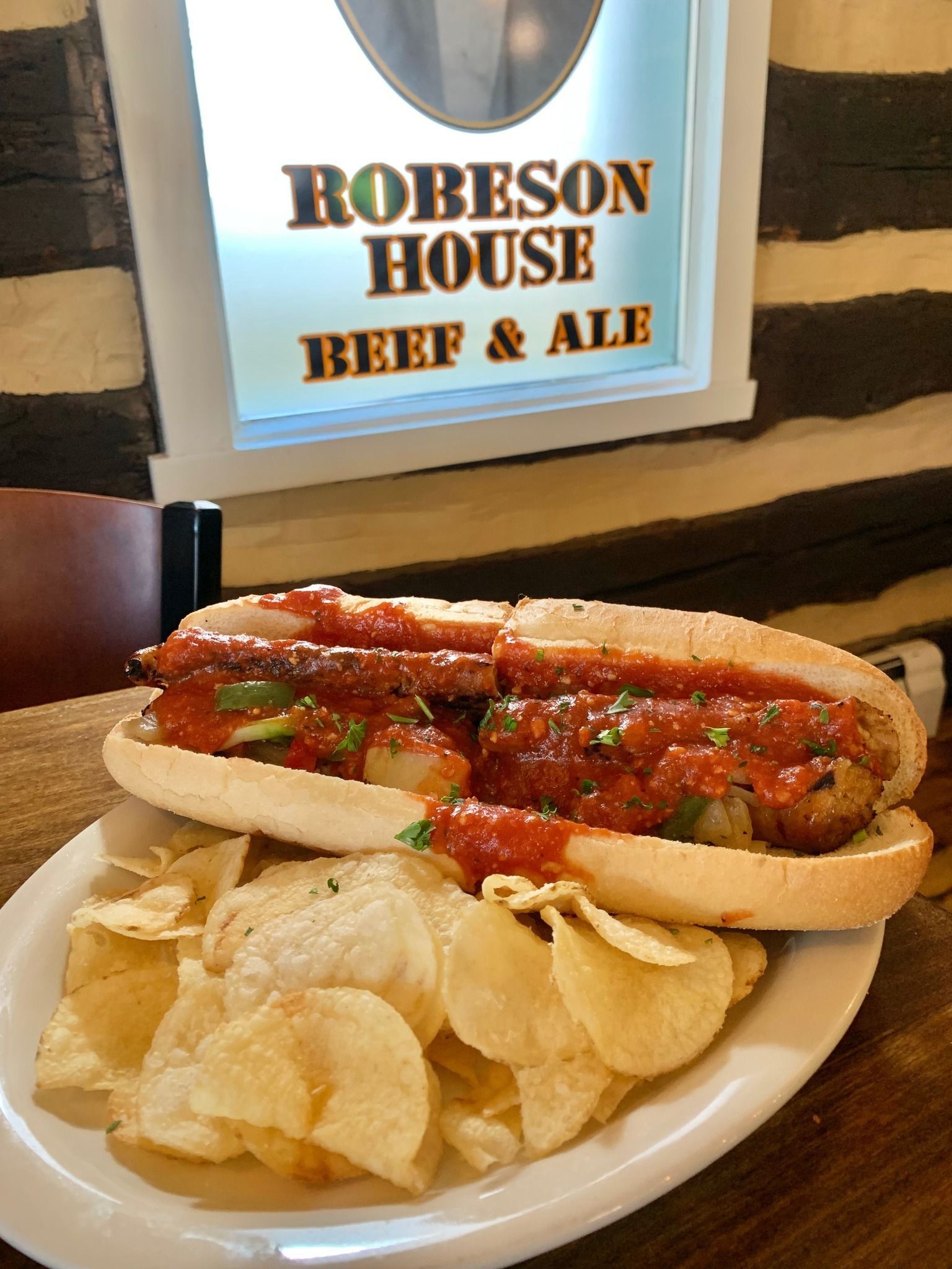A hot dog and chips are on a plate in front of a sign that says robeson house beef & ale