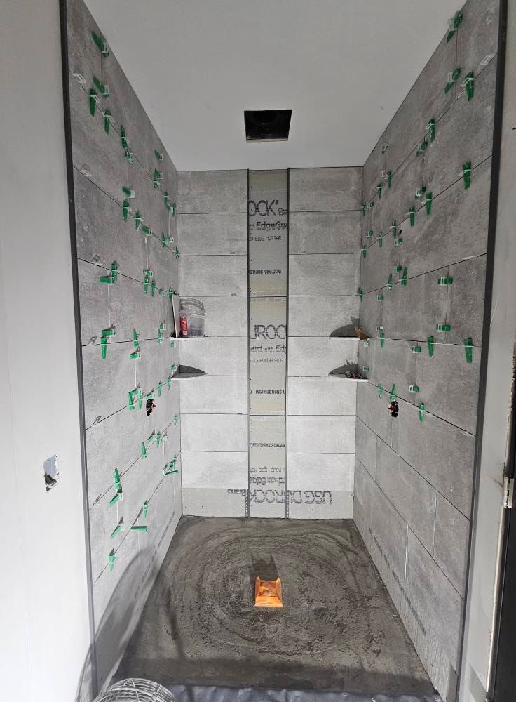Shower stall under construction, gray tiled walls with green spacers, shelves, and a drain.