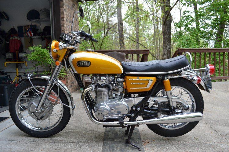 Full left-side view of the 1971 yamaha xs1b. Has trees as a back drop and very corner of garage and fence railing running left to right
