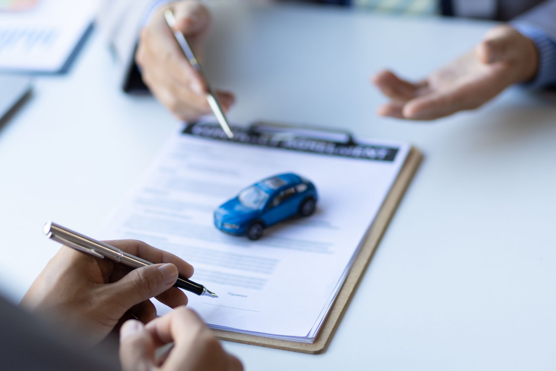 Close-up of a hand signing a contract with a blue model car on top, from an auto insurance provider. Close-up of a hand signing a contract with a blue model car on top, from an auto insurance provider.
