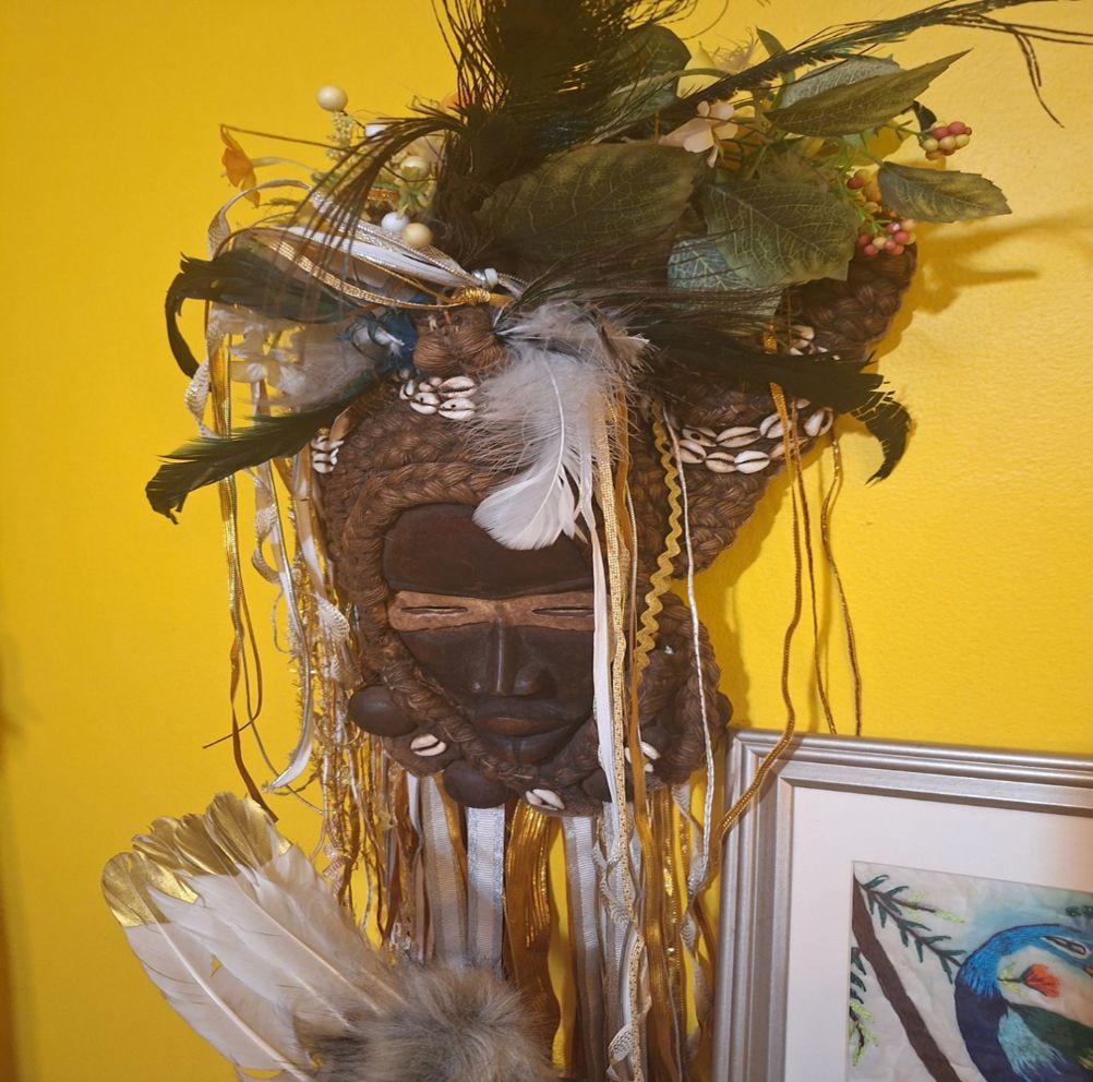 Mask with feathers, shells, and foliage against a yellow wall; a picture frame is below.