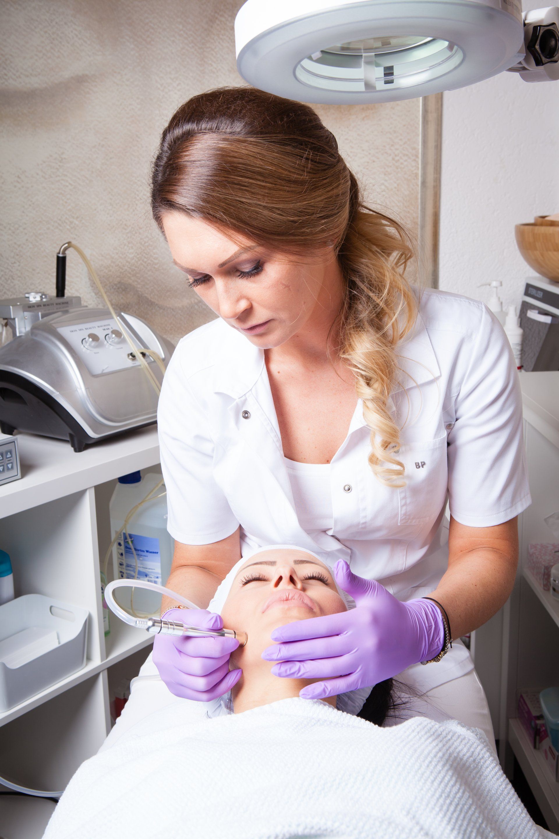 Eine Frau mit lila Handschuhen reinigt in einem Schönheitssalon das Gesicht einer anderen Frau.