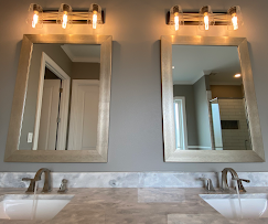 Two bathroom mirrors with silver frames and matching light fixtures above a marble countertop with sinks and faucets.