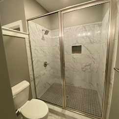 Bathroom with a marble-tiled shower behind glass doors. Toilet to the left, and a shower head is visible.