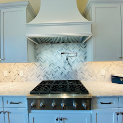 A kitchen stove and hood with blue-grey cabinets, white marble countertops, and a herringbone tile backsplash.