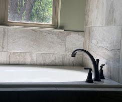 Bathtub with black faucet against gray tile wall, window in background.
