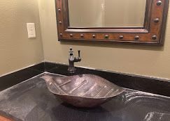 Copper leaf-shaped sink on black countertop, under a brown framed mirror in a bathroom.
