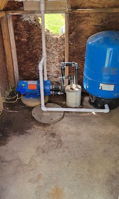 Well water pump system with a blue pressure tank and pump, PVC pipes, and a well cap in a small shed.