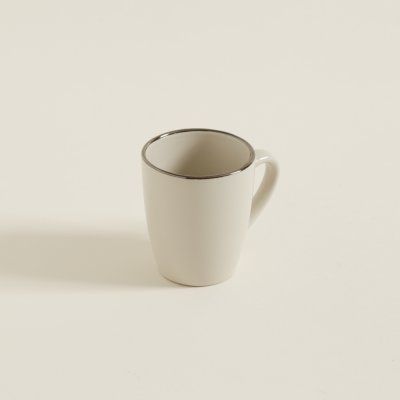 Una taza de café blanca con borde plateado reposa sobre una superficie blanca.