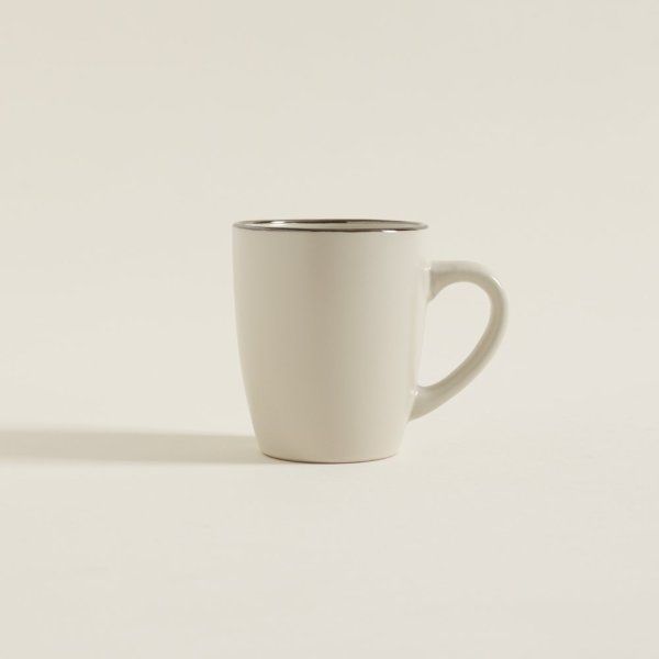 Una taza de café blanca con borde plateado reposa sobre una superficie blanca.