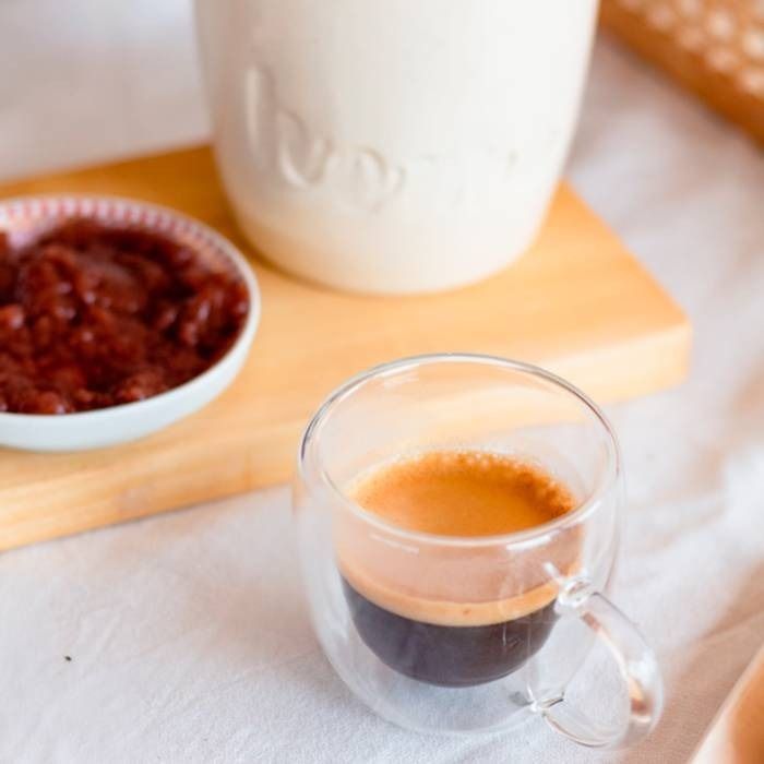 Una taza de café reposa sobre una mesa junto a un tazón de mermelada.