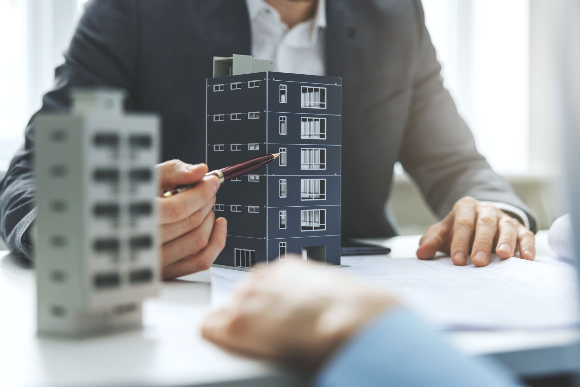 Man in suit pointing at a model building, discussing with another person at a desk.