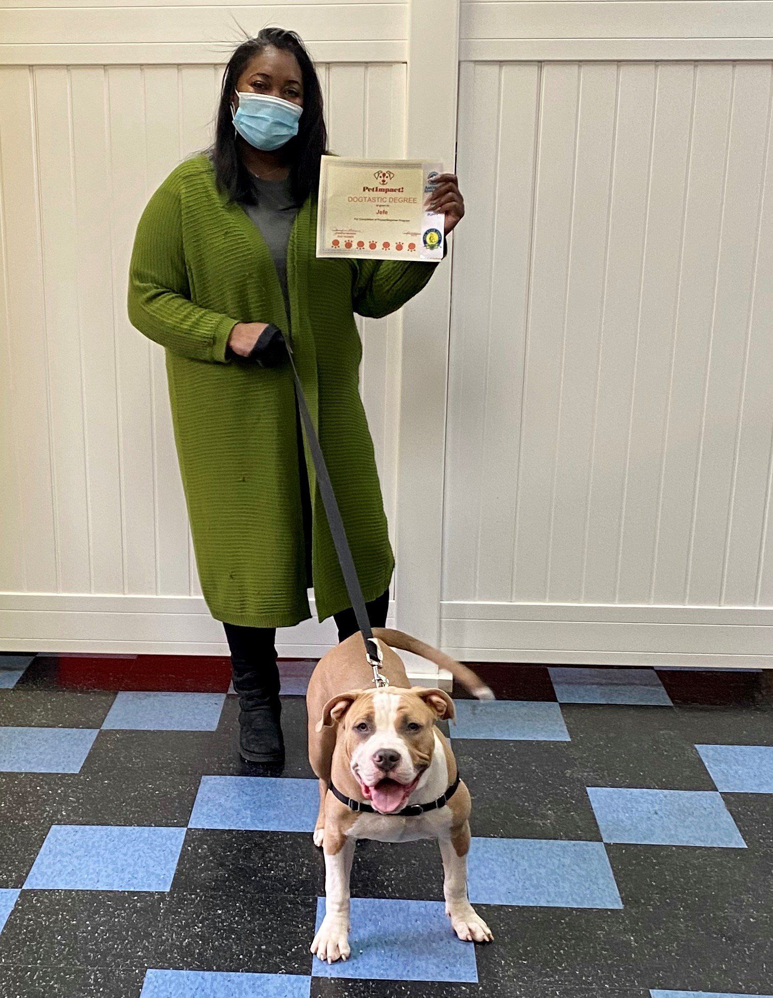 Woman in green sweater and a dog holding a certificate. Blue and black checkered floor, white walls.