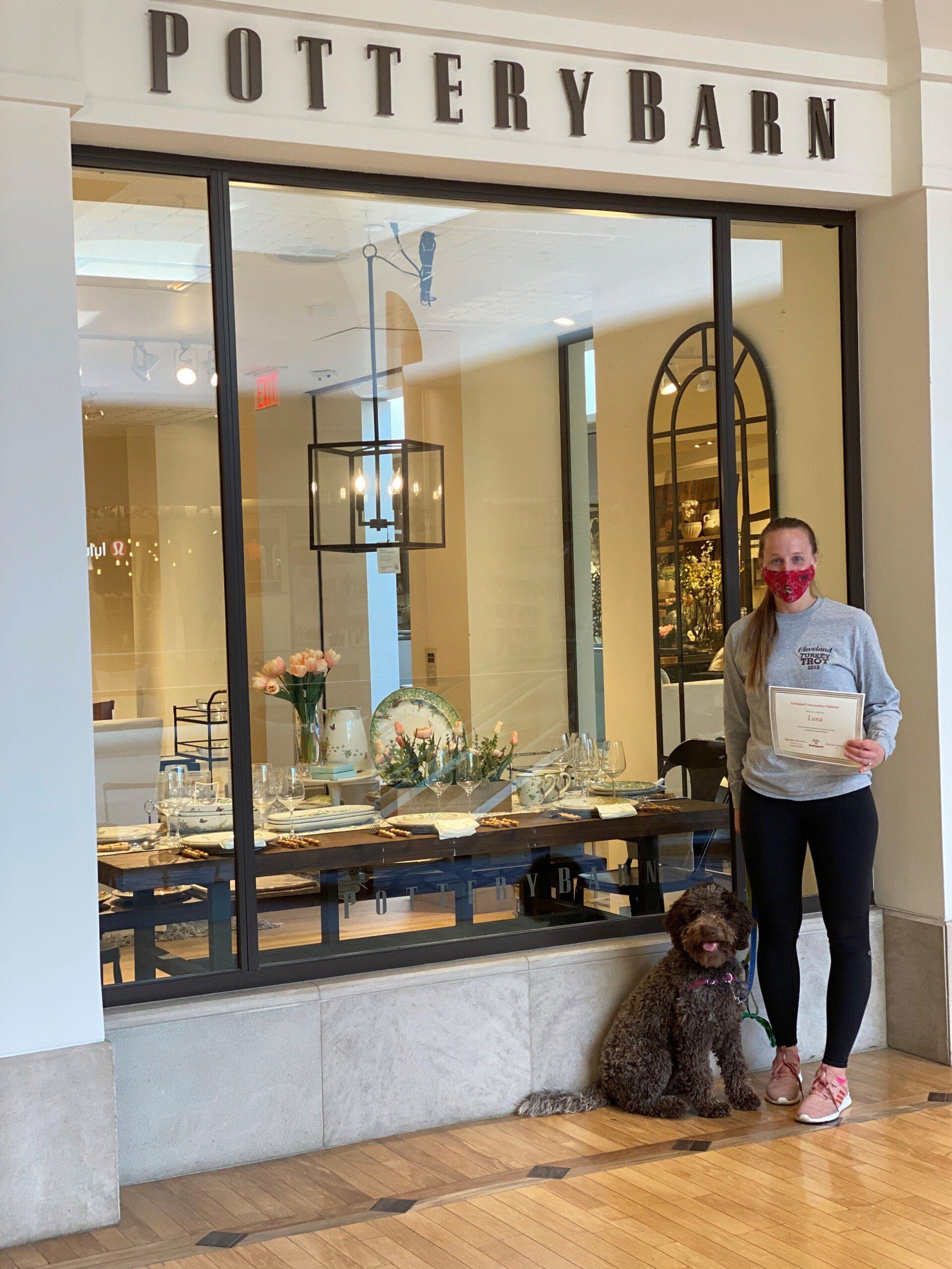 Woman and dog pose in front of a Pottery Barn window display. Woman wears a mask and holds a certificate.