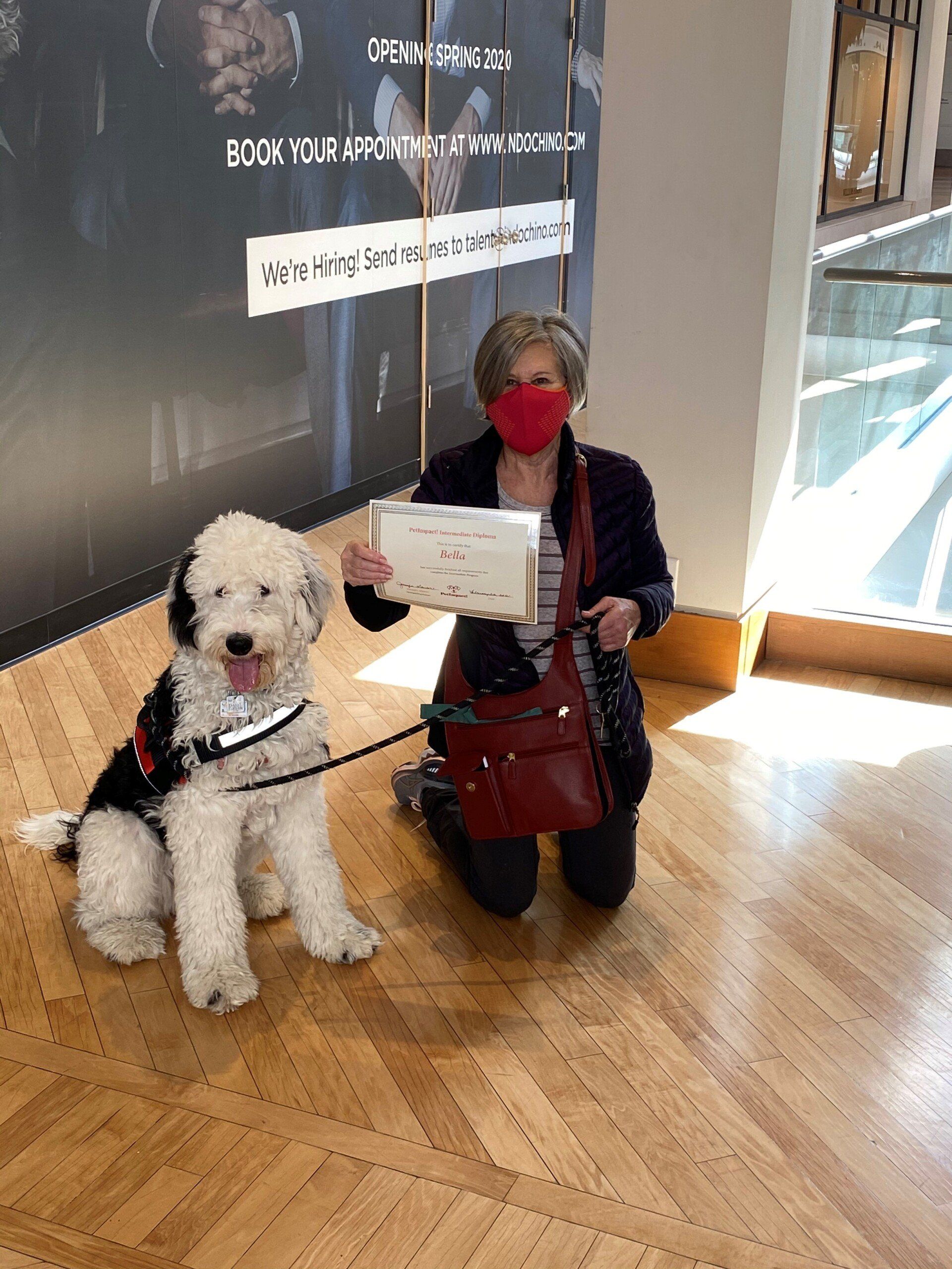 Woman kneeling with a service dog; both looking at the camera. Woman holds a paper; wall with text behind them.