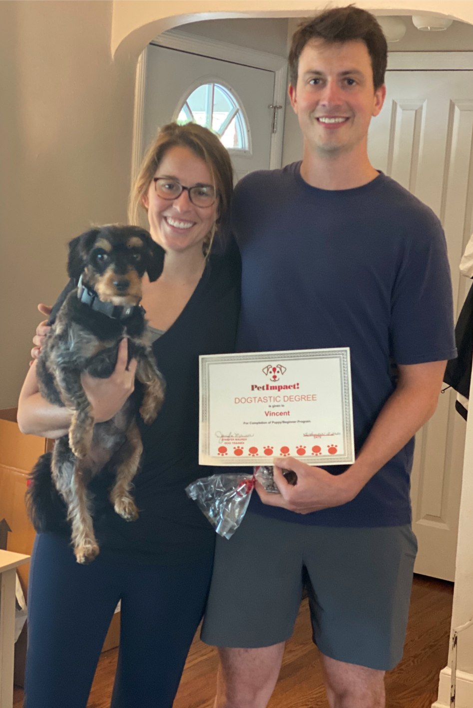 Couple holds a certificate and dog, smiling. Indoors, neutral walls, natural light.