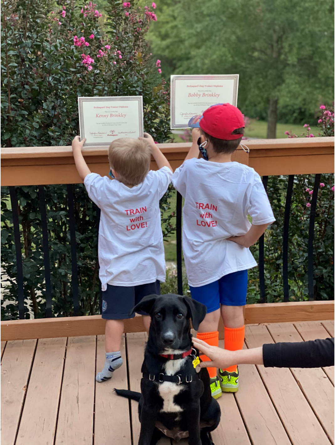 Two children holding certificates on a deck, dog sits at their feet. The children wear white t-shirts.