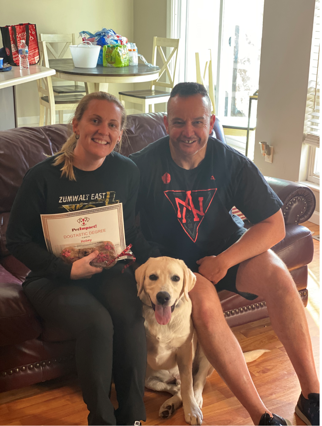 Woman and man sit with a yellow lab on a brown couch; woman holds a certificate.