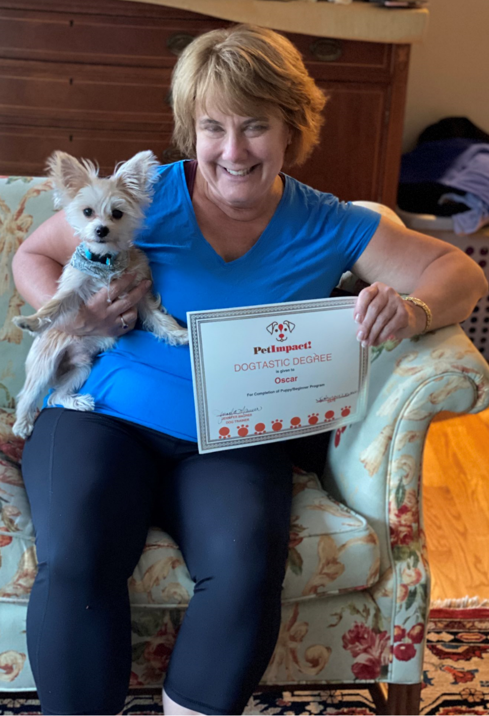 Woman with dog, holding a certificate. She is wearing blue top, black pants, sitting on floral chair.
