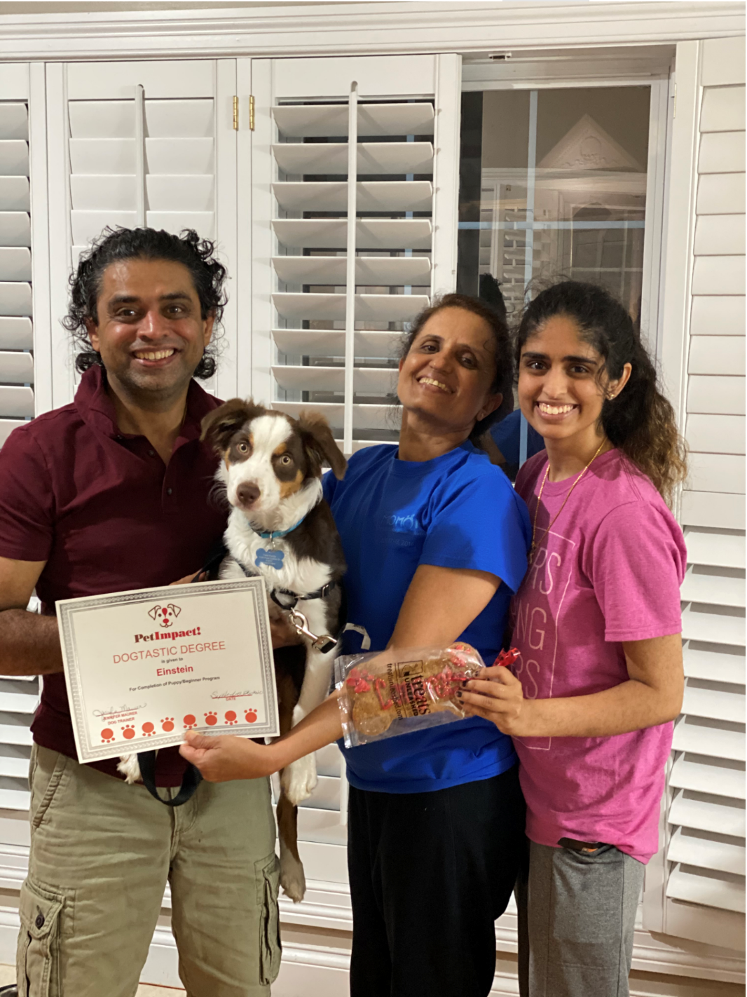 Three people and a dog holding a certificate and treats in front of a window. They are smiling.