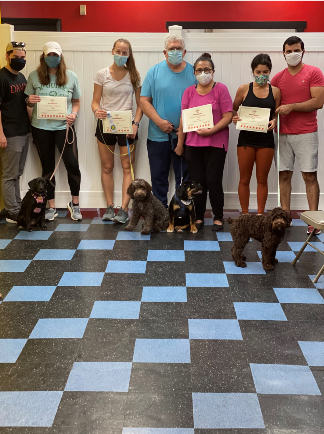 Group of people and dogs holding certificates; posing in a room with checkered floor.