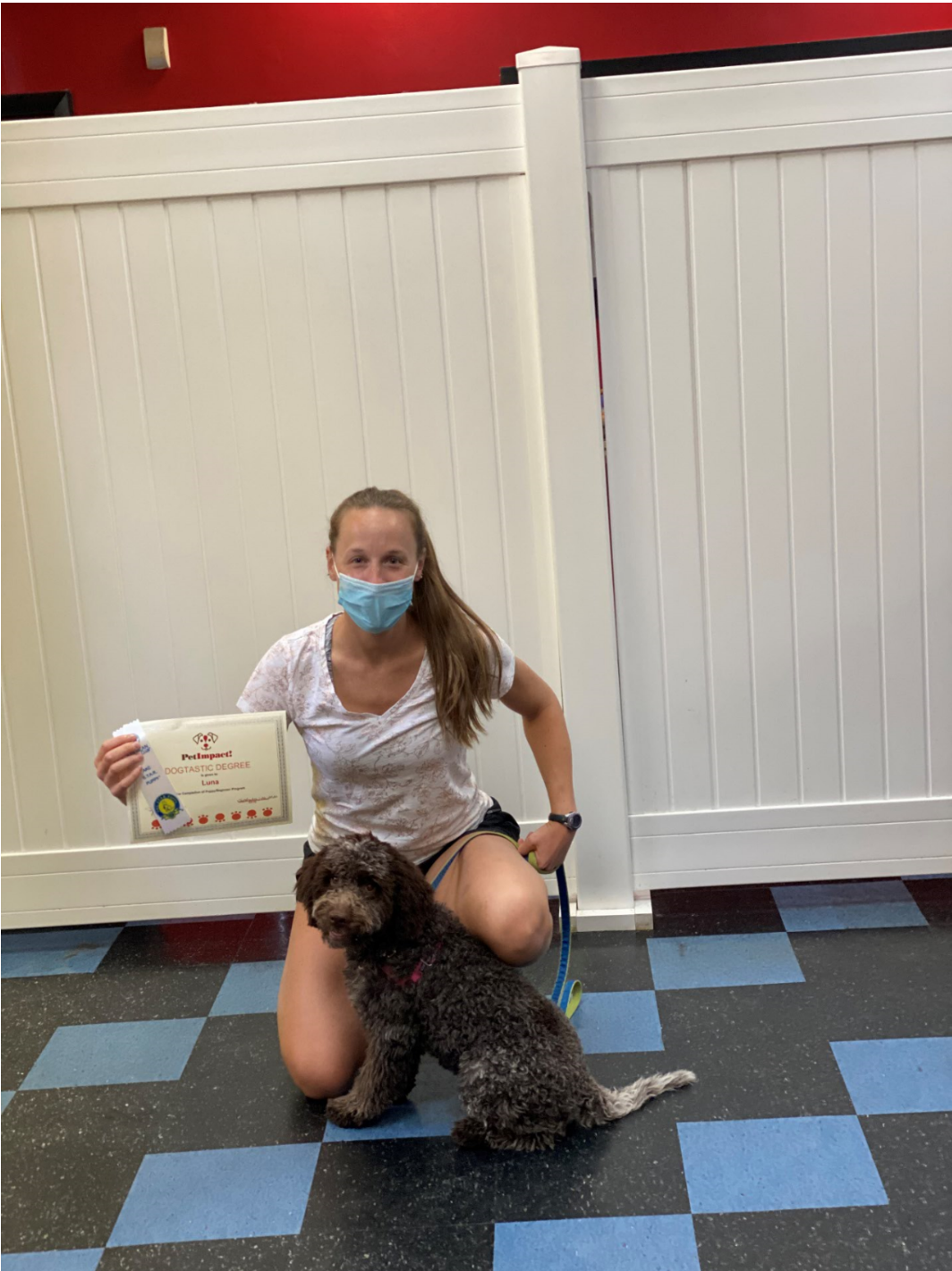 Woman in mask kneels with dog, holding certificate. They are in a room with a white fence background and blue tiled floor.