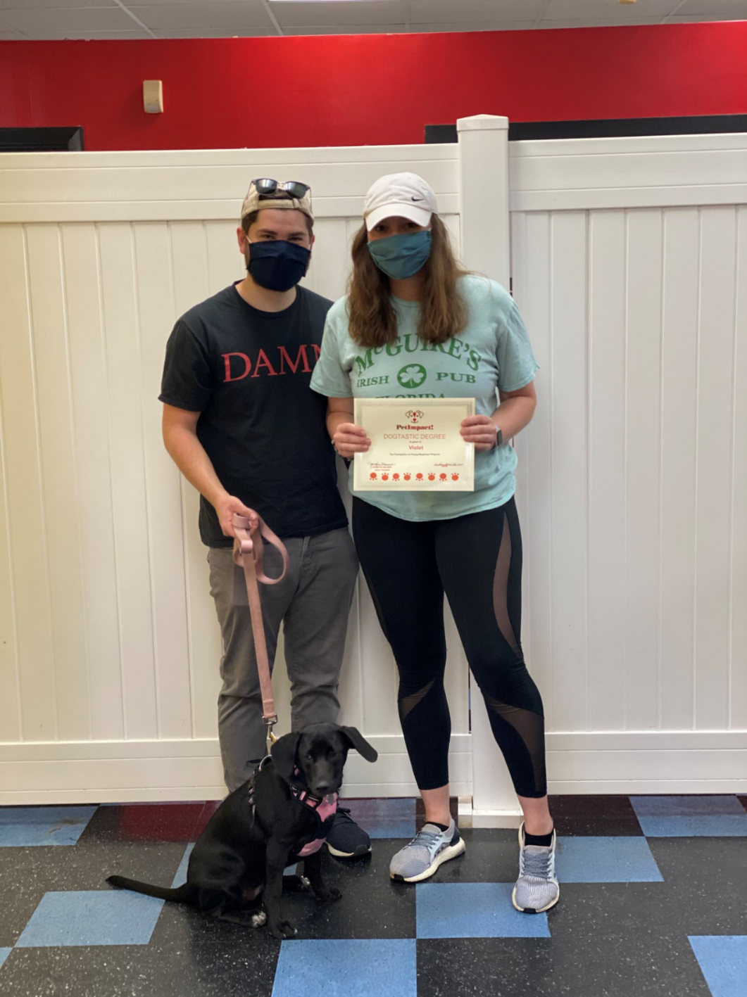 People and dog pose with certificate in front of a white wall and a blue floor.