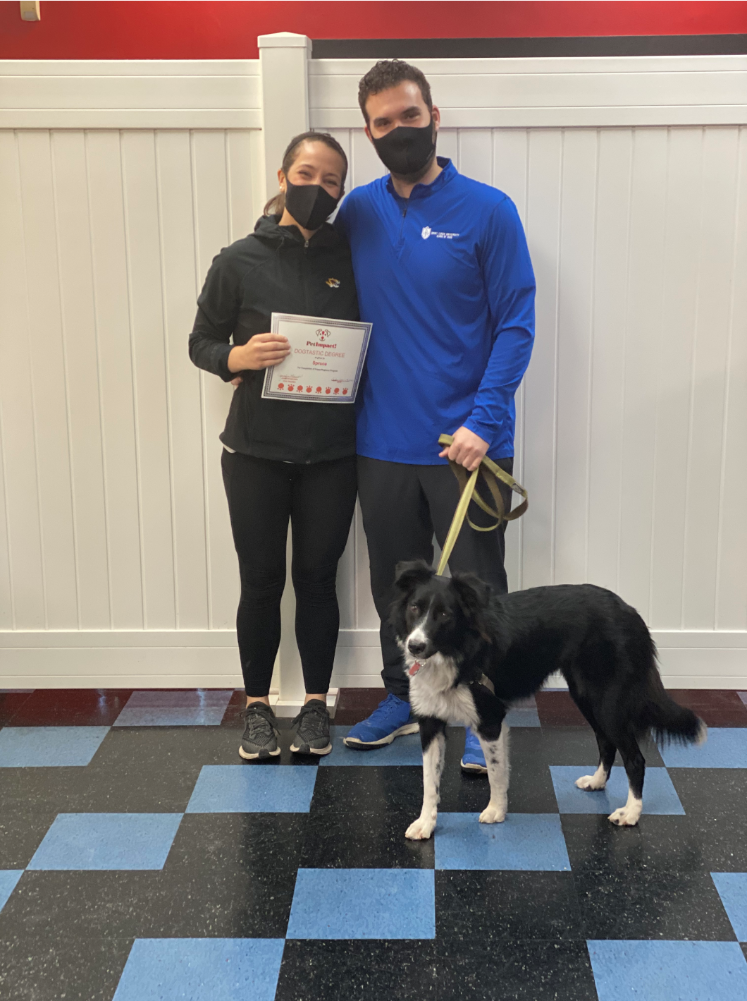 A woman and man with a border collie dog. Woman holds a certificate; all wear masks in a gym.