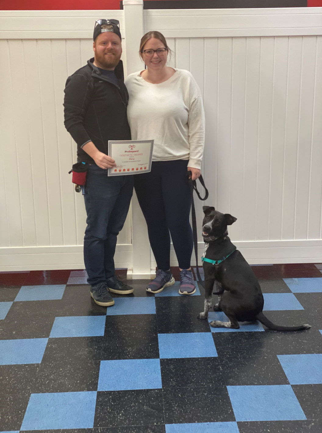 Couple and dog pose with a certificate. They stand in front of a white fence. Dog sits.