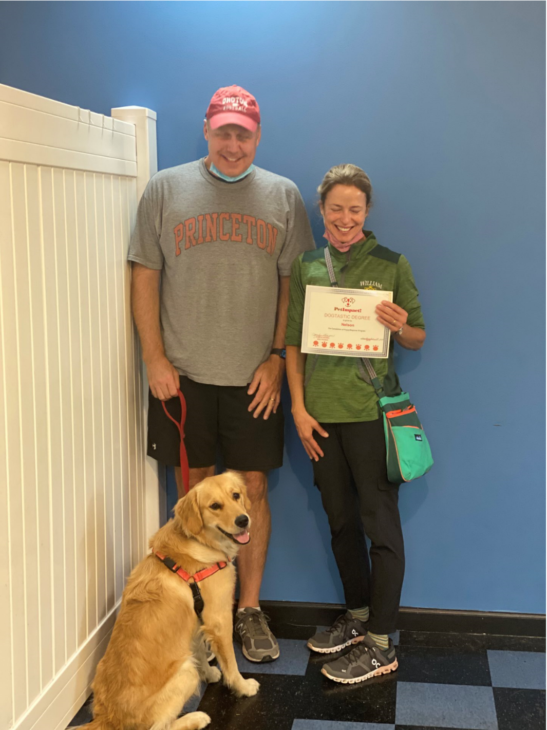 A golden retriever sits with a man and woman holding a certificate. Blue wall background.