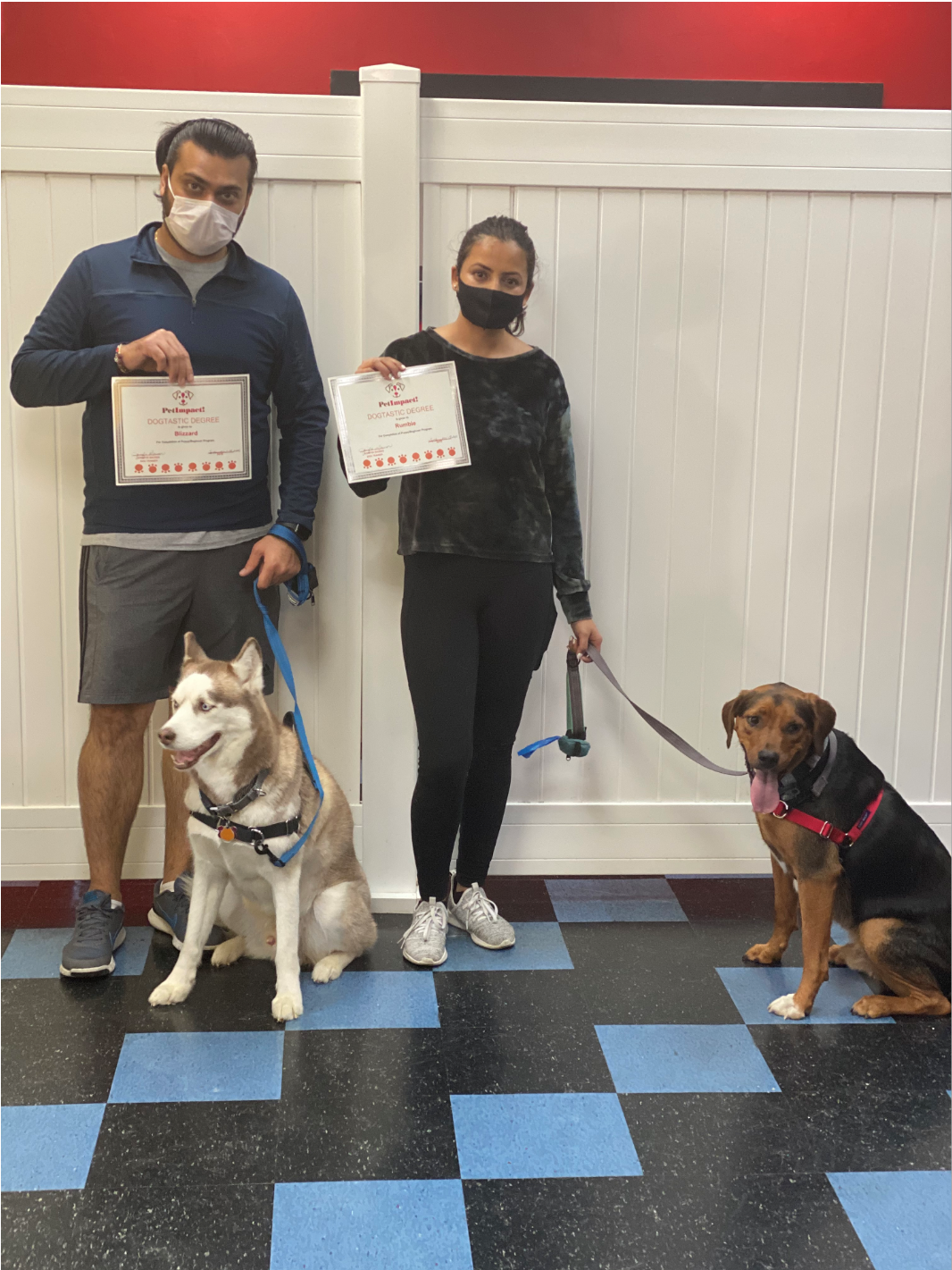 Two people with dogs hold certificates; they stand indoors with a red-white wall background and blue flooring.