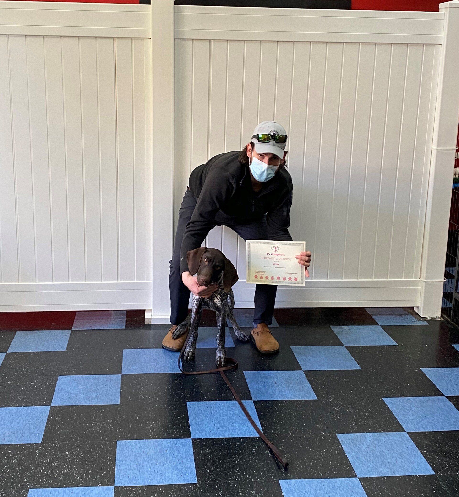 Man in mask holds dog and certificate near checkered floor. White fence backdrop.