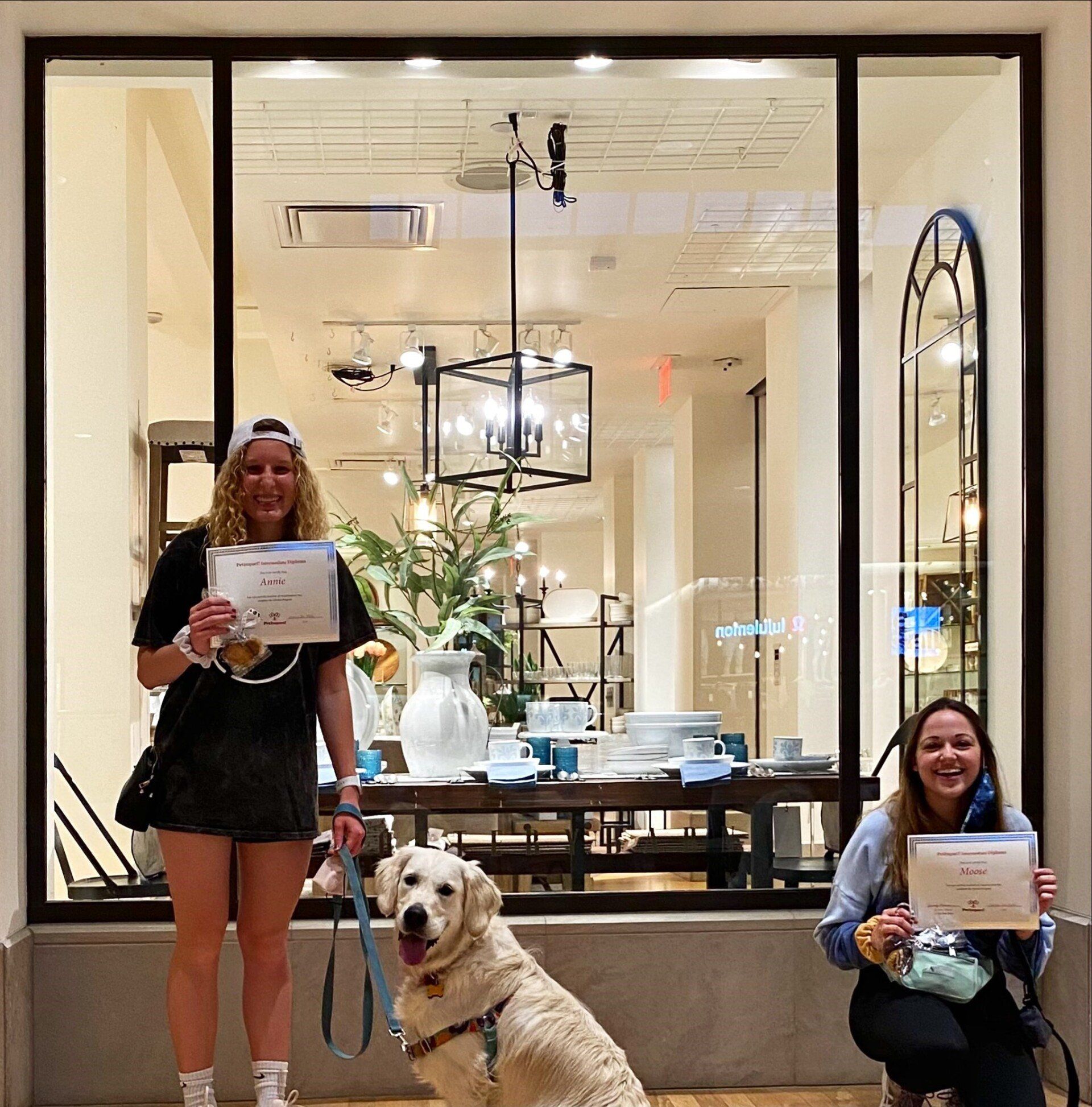 Two women and a golden retriever pose outside a store window, holding certificates.