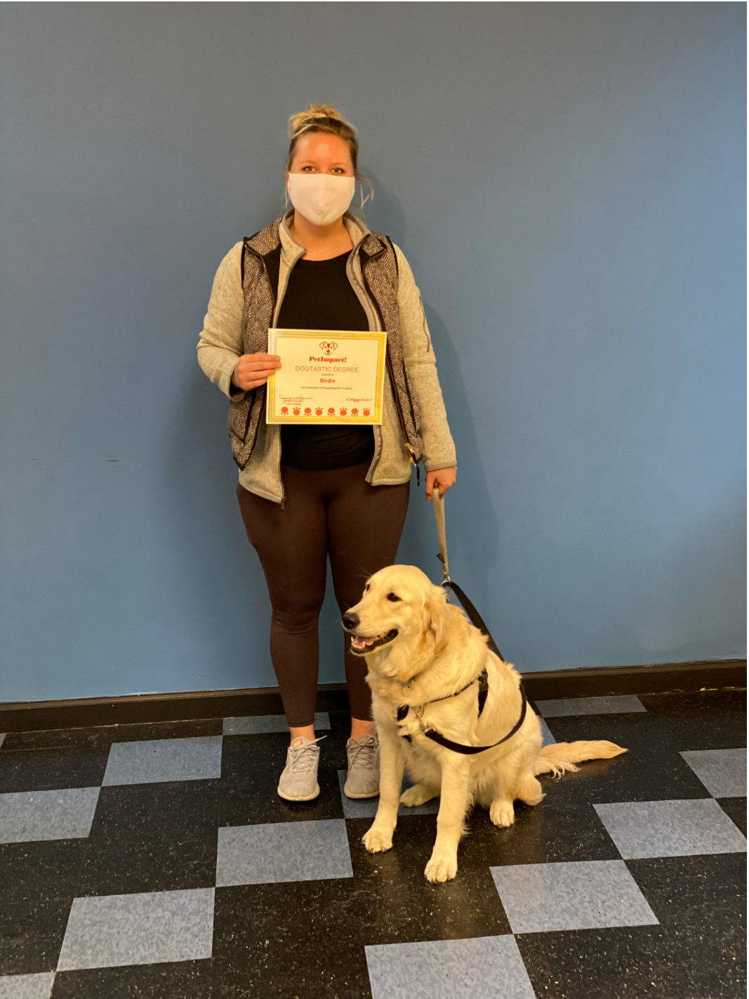 Woman with service dog, holding certificate, in front of a blue wall. Black and gray checkered floor.