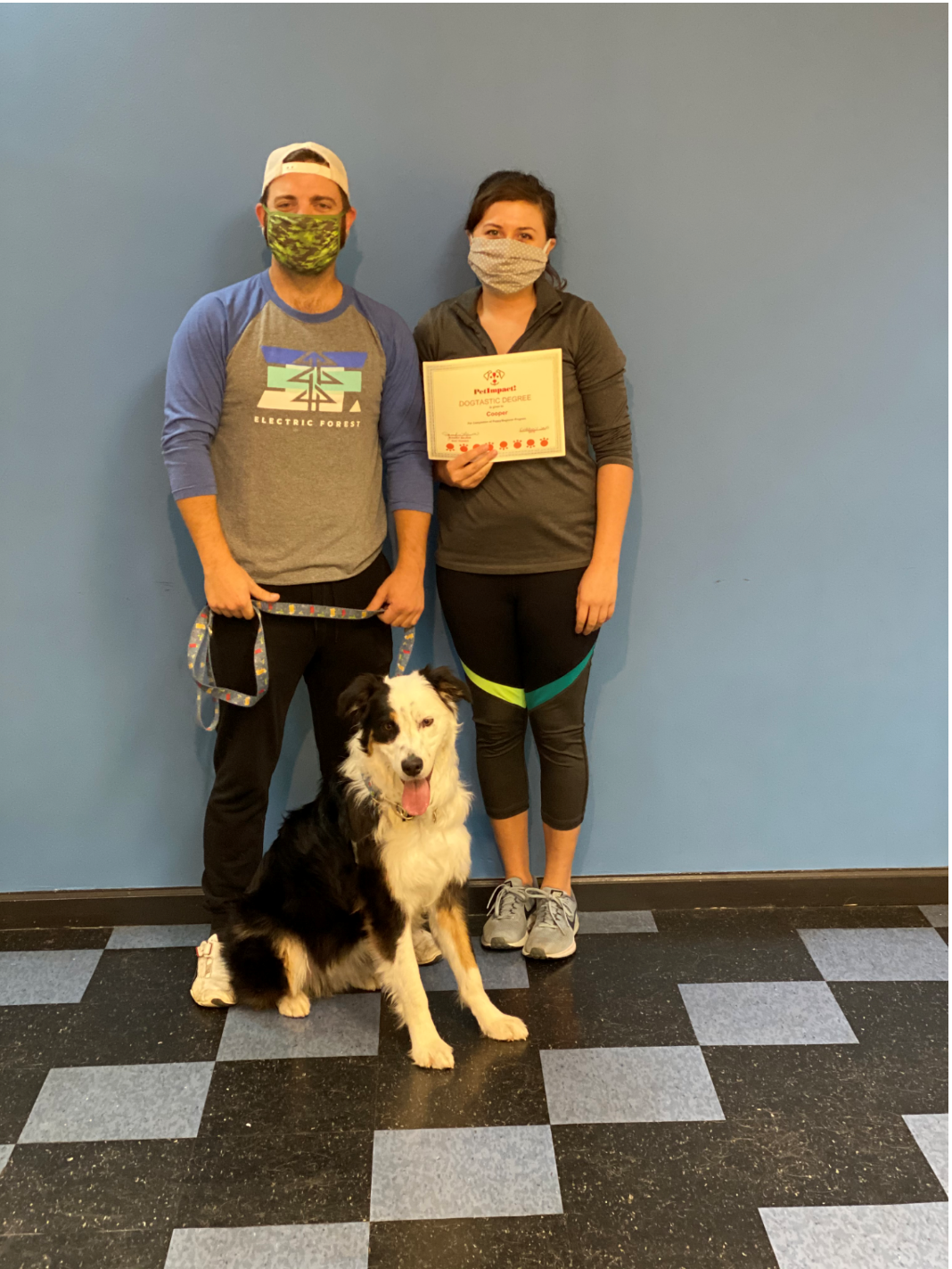 A dog sits with two people, holding a certificate, posing in front of a blue wall.