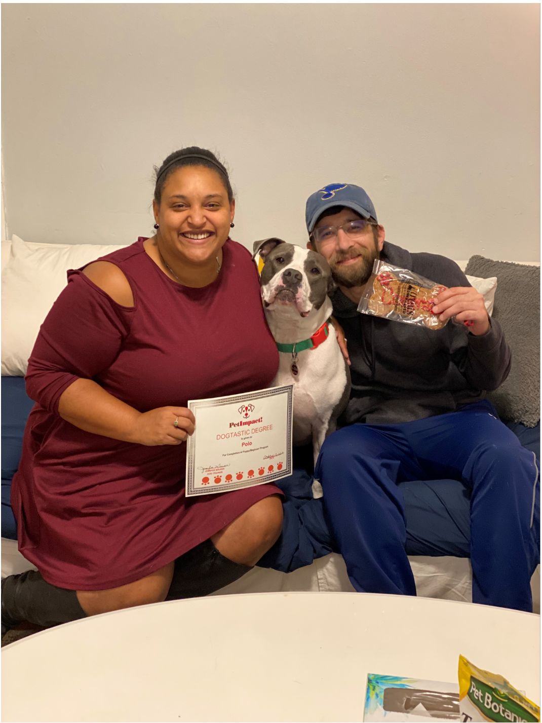 Couple and dog pose on couch, holding award and treats. Woman in burgundy dress, man in blue hat.