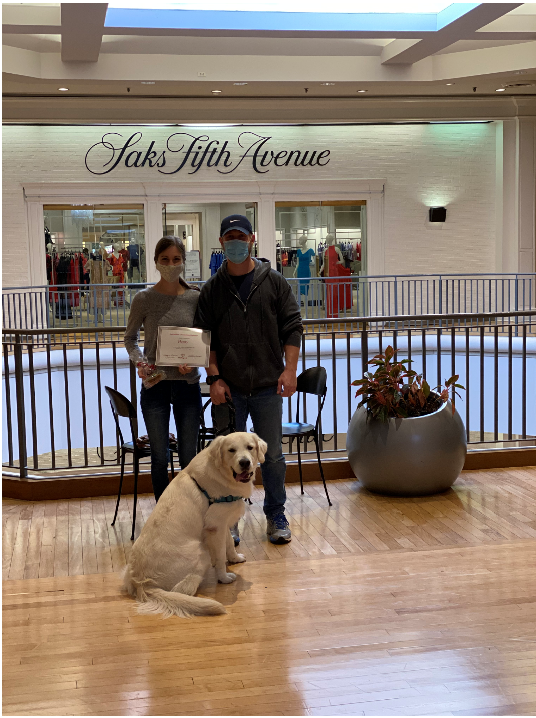 A dog sits with two people in front of Saks Fifth Avenue. People wear masks, one holds a paper.