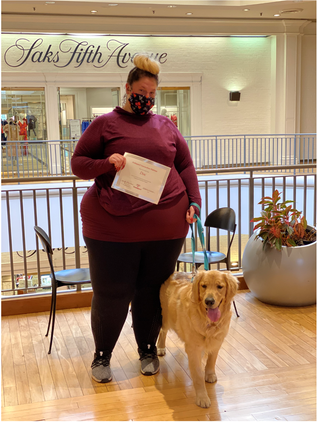Woman in maroon shirt and black pants with golden retriever, holding a paper in front of Saks Fifth Avenue.